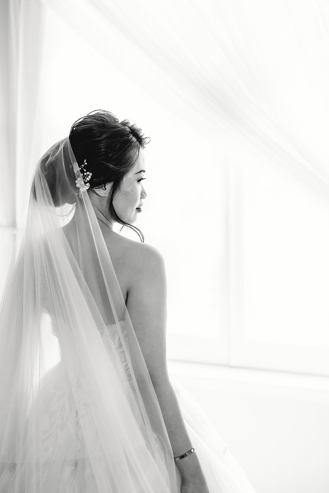 A bride in a veil looking towards the window, photographed in black and white